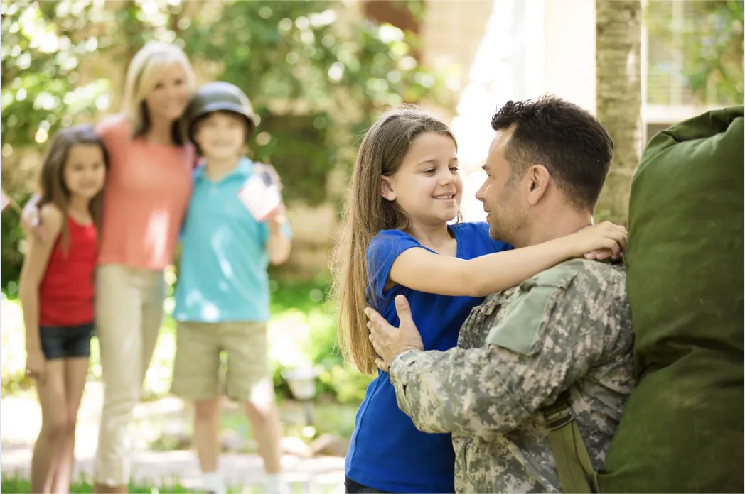 Military parent embracing daughter; family blurred in background with flag